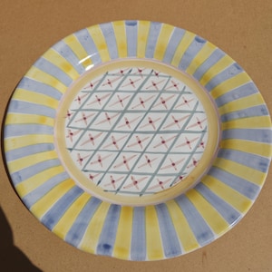 May include: A round ceramic plate with a yellow and blue striped border. The center of the plate features a white background with a crisscross pattern and small red star-like shapes. The plate has a yellow inner ring.