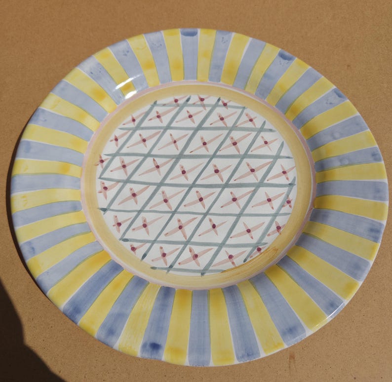 May include: A round ceramic plate with a yellow and blue striped border. The center of the plate features a white background with a crisscross pattern and small red star-like shapes. The plate has a yellow inner ring.