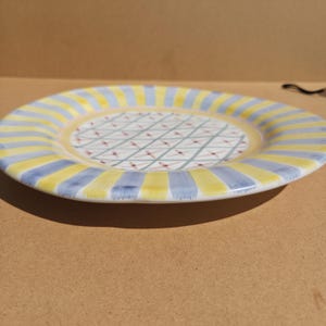 May include: A ceramic plate with a yellow and blue striped rim. The center of the plate has a white background with a crisscross pattern of red and green lines. The plate is round and has a slightly raised edge.