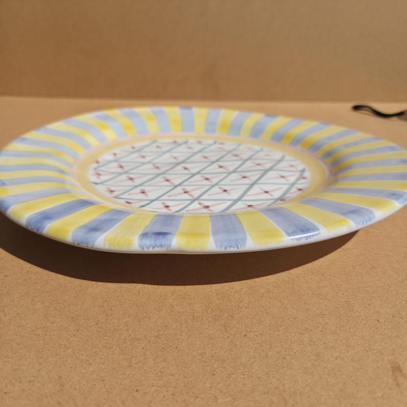 May include: A ceramic plate with a yellow and blue striped rim. The center of the plate has a white background with a crisscross pattern of red and green lines. The plate is round and has a slightly raised edge.
