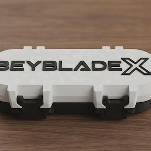 Pode incluir: Um estojo de armazenamento oblongo, branco e preto, com um padrão geométrico texturizado na tampa. O topo do estojo apresenta o texto BEYBLADEX em relevo preto. O recipiente possui dois fechos pretos na frente e um formato arredondado.