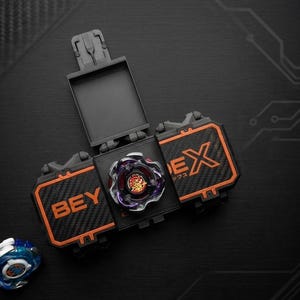 K&ouml;nnte beinhalten: Ein schwarzes und orangefarbenes Beyblade-Spielzeugset mit einem Kreisel in der Mitte. Das Set enth&auml;lt einen Werfer und einen Ersatzkreisel. Die W&ouml;rter "BEY" und "EX" sind auf dem Set sichtbar.
