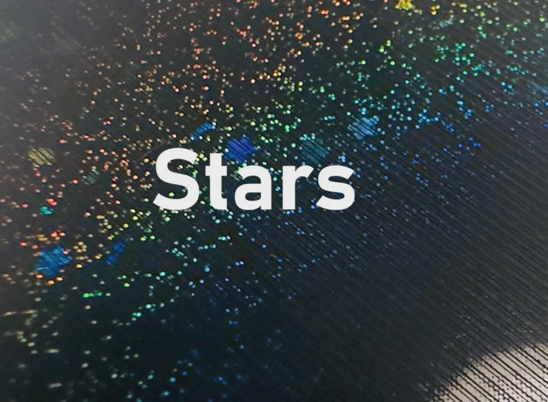 K&ouml;nnte beinhalten: Abstraktes Bild mit dem Wort "Stars" in Wei&szlig;. Der Hintergrund ist dunkel und mit kleinen, bunten, irisierenden Punkten bedeckt, die an einen Sternenhimmel erinnern. Das Design hat eine schimmernde, himmlische Qualit&auml;t.