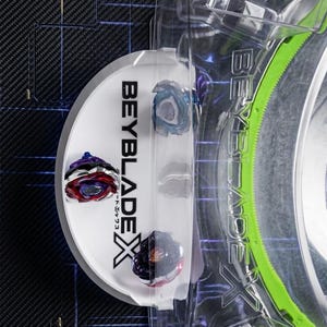 Könnte beinhalten: Eine transparente Kunststoffverpackung mit drei farbigen Beyblade X-Kreiseln. Die Verpackung hat das Beyblade X-Logo in Schwarz und Weiß. Die Kreisel sind in verschiedenen Farben gehalten, darunter Rot, Blau und Schwarz. Die Verpackung befindet sich auf einer strukturierten schwarzen Oberfläche.