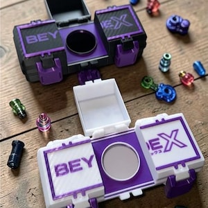 Könnte beinhalten: Zwei offene, lila und weiße Spielzeugaufbewahrungsboxen mit den Aufdrucken "BEY" und "EX". Verschiedene bunte Spielzeugteile sind auf einer Holzoberfläche um die Boxen herum verstreut.