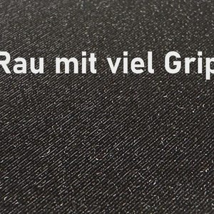 K&ouml;nnte beinhalten: Nahaufnahme einer strukturierten schwarzen Oberfl&auml;che mit dem wei&szlig;en Text "Rau mit viel Grip". Die Oberfl&auml;che hat ein diagonales Muster und kleine wei&szlig;e Flecken. Der Text bedeutet "Rau mit viel Grip".