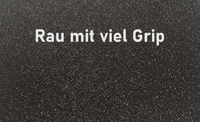 K&ouml;nnte beinhalten: Nahaufnahme einer strukturierten schwarzen Oberfl&auml;che mit dem wei&szlig;en Text "Rau mit viel Grip". Die Oberfl&auml;che hat ein diagonales Muster und kleine wei&szlig;e Flecken. Der Text bedeutet "Rau mit viel Grip".