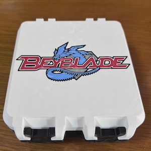 Può includere: Una custodia rettangolare in plastica bianca con il logo Beyblade rosso e nero e un grafico di un drago blu centrato sul coperchio. La custodia presenta due chiusure nere sul bordo anteriore e una finitura superficiale bianca liscia.