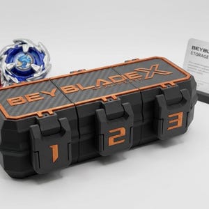 K&ouml;nnte beinhalten: Schwarze und orangefarbene Beyblade X Aufbewahrungsbox mit drei nummerierten F&auml;chern. Zwei Beyblades, eines rot und gold, das andere blau und silber, sind sichtbar. Eine Zubeh&ouml;rkartenkarte ist ebenfalls vorhanden.