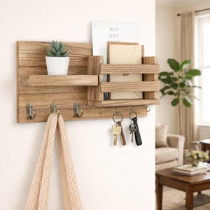May include: Wooden wall-mounted organizer with a shelf, mail slots, and key hooks. It features a small potted succulent, a card that reads "JUST LOVE", and hanging keys. A beige scarf hangs from one of the hooks.
