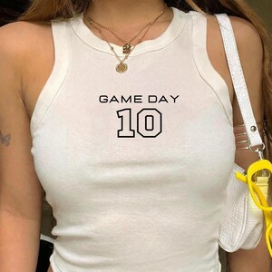 Op de afbeelding: Witte tanktop met de tekst "GAME DAY" en het zwarte nummer "10". De top is mouwloos en geribbeld. Een witte handtas met een witte riem en gele zonnebril zijn ook zichtbaar.