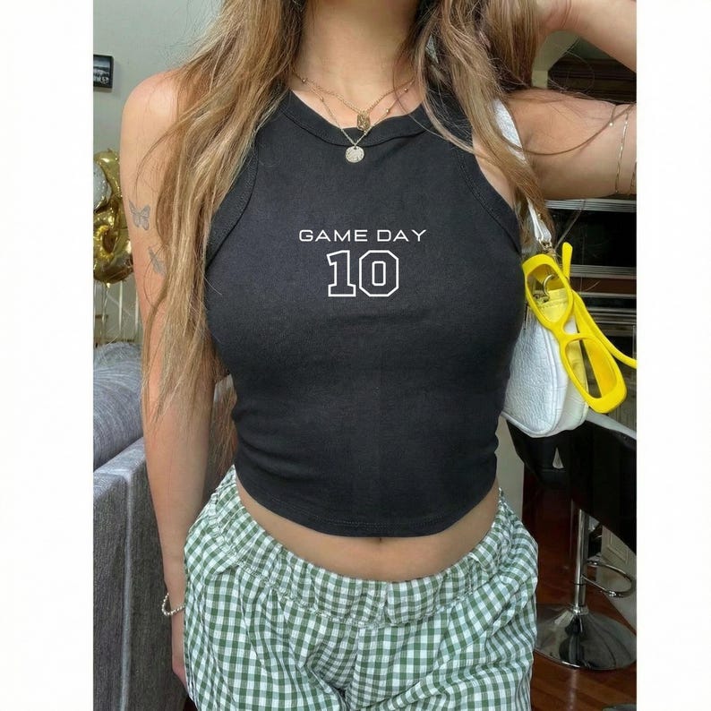 Op de afbeelding: Zwart geribbelde tanktop met de tekst "GAME DAY 10" in het wit. De top wordt gecombineerd met een groen-witte geruite short. Een witte handtas en gele zonnebril zijn ook zichtbaar.