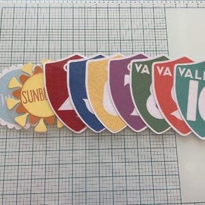 May include: A collection of die-cut paper shapes including a yellow sun with the text SUNBEAM and several shield-shaped cards in red, blue, yellow, purple, and green, featuring the text VALIANT and numbers ranging from 7 to 10 in white font.