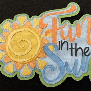 May include: A layered paper die-cut design with a sun and the words "Fun in the Sun". The sun is yellow and orange, and the text is orange and blue. The design has a green border and is on a black background.