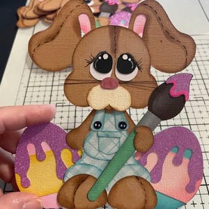 May include: A brown bunny-shaped paper cutout holding a paintbrush, with two painted Easter eggs. The bunny wears blue overalls and has large, expressive eyes. The eggs are decorated with purple and pink paint. Additional bunny cutouts are in the background.
