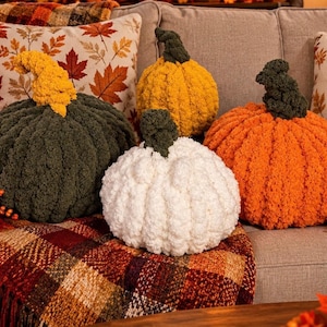 May include: A collection of four handmade chunky knit pumpkin pillows in dark green, mustard yellow, white, and orange. Each pumpkin features a textured, soft yarn construction with a distinct stem in a contrasting color, arranged together as decor.