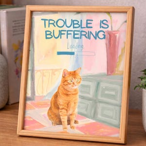 Può includere: Una stampa incorniciata con una cornice in legno raffigurante un'illustrazione di un gatto rosso. L'opera d'arte include il testo "TROUBLE IS BUFFERING Loading" in blu. Il gatto è seduto di fronte a uno sfondo colorato con un vaso rosa.