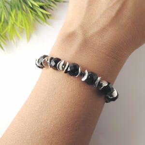 Op de afbeelding: Een elastische armband met afwisselend ronde, gefacetteerde zwarte kralen en kleine, getextureerde zilverkleurige metalen tussenstukjes. De kralen zijn in een herhalend patroon gerangschikt en vormen een doorlopend cirkelvormig ontwerp.