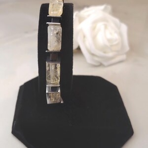 May include: A bracelet with rectangular, pale yellow gemstones set in silver metal. The bracelet is displayed on a black velvet stand, with a white rose in the background. The gemstones have a translucent appearance.