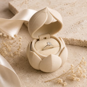May include: A cream-colored ring box shaped like a lotus flower with a hinged lid, containing a silver ring with a single round-cut clear stone. The interior of the box is lined with soft, light-colored fabric to hold the ring securely in place.