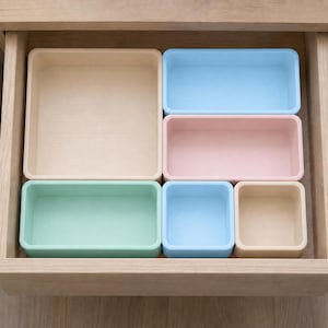 May include: A set of six rectangular and square storage organizers in various pastel colors including beige, light blue, soft pink, and mint green. The containers are arranged to fit together within a wooden drawer, featuring smooth edges.