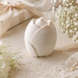 May include: A small, off-white, egg-shaped container designed in the likeness of a closed flower bud. The object features smooth, overlapping petal-like segments that form a rounded exterior, with a horizontal seam indicating a lid that opens.