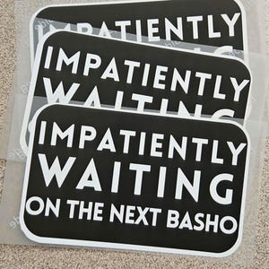 Può includere: Tre adesivi rettangolari neri con testo bianco "IMPATIENTLY WAITING ON THE NEXT BASHO". Gli adesivi hanno angoli arrotondati e un bordo bianco.