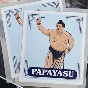 Puede incluir: Dos pegatinas con una ilustración de dibujos animados de un luchador de sumo con un moño negro, vistiendo un mawashi azul. Las pegatinas tienen un fondo azul claro con la palabra "PAPAYASU" en blanco sobre una pancarta azul.
