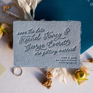 May include: A light blue wedding invitation with the text "save the date Rachel Nancy & George Everett are getting married June 3, 2026". The invitation is surrounded by dried flowers and a teal envelope.