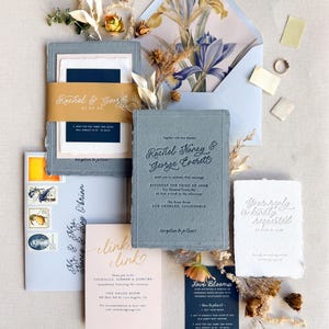 May include: Wedding invitation suite featuring a blue-grey invitation, RSVP, and details cards. The suite includes a floral-lined envelope, a gold belly band, and a small navy card. Dried flowers and a ring are also included.