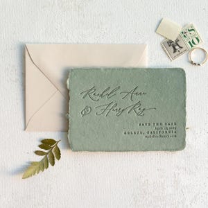 May include: A sage green wedding invitation with the names "Rachel Anne & Henry Ray" in elegant script. The text "Save the Date" and wedding details are also included. A cream envelope, stamps, and a ring are also visible.