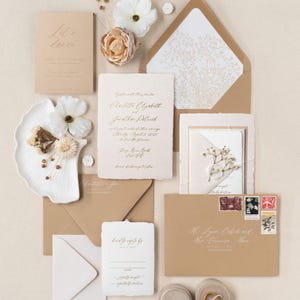 May include: An assortment of wedding stationery, including invitations, reply cards, and envelopes, arranged on a neutral surface. The invitations feature elegant gold calligraphy. Decorative flowers and stamps add to the aesthetic.