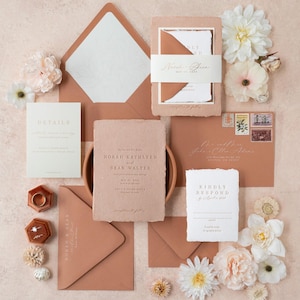 May include: A flat lay of wedding stationery in shades of peach and white. The set includes invitations, envelopes, and response cards, with floral accents and gold text. The invitation reads "Norah Kathleen and Sean Walter".