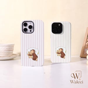 May include: Two smartphone cases featuring a light pink and white vertical striped pattern. Each case displays a central graphic of a brown monkey character sleeping on a white pillow while hugging a small tan teddy bear, with black camera cutouts.