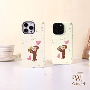 May include: Two cream-coloured phone cases featuring a brown cartoon monkey holding a small circular mirror. The design includes pink hand-drawn hearts and yellow stars scattered across the surface. Each case has a black camera lens cutout.