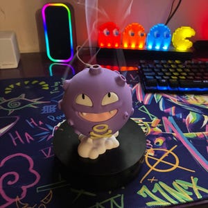 Pode incluir: Um queimador de incenso esférico roxo em forma de personagem de desenho animado com um sorriso largo, dois dentes visíveis e um símbolo amarelo de caveira e ossos cruzados no peito. A figura assenta numa base de nuvem branca e emite fumo.