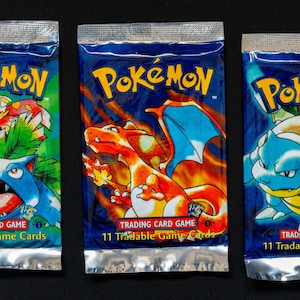 May include: Three sealed Pokémon trading card packs. Each pack features a different Pokémon character illustration in vibrant colors. The packs have a blue background with the Pokémon logo and text that reads "Trading Card Game" and "11 Tradable Game Cards".