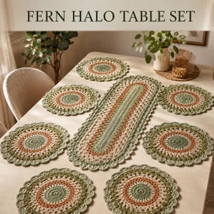 May include: A Fern Halo Table Set consisting of one oval crochet runner and six circular crochet placemats. Each piece features concentric rings of sage green, terracotta, and cream yarn with a scalloped edge. The text reads FERN HALO TABLE SET.