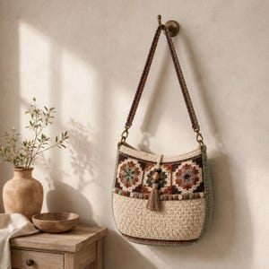 Puede incluir: Un bolso de hombro de ganchillo con un patrón de cuadrados de la abuela multicolor en tonos marrón, verde y crema. El bolso tiene una correa larga estampada, un detalle de cuenta de madera central y una borla decorativa en el centro.