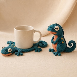 Puede incluir: Un posavasos de ganchillo con forma de lagarto en color verde azulado y melocotón con una taza de cerámica blanca encima, acompañado de una figura de lagarto de ganchillo a juego de pie con una cola enroscada y pequeñas manchas amarillas.