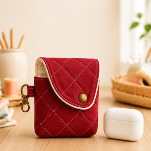 Peut inclure: Une pochette en tissu rouge foncé matelassé avec un motif en losanges, un rabat arrondi et un bouton-pression en laiton. Un mousqueton en métal couleur laiton est fixé sur le côté. Un boîtier de charge blanc pour écouteurs est posé à côté.