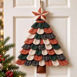 May include: A fabric wall hanging shaped like a Christmas tree, featuring layered, scalloped felt pieces in shades of dark green, cream, and rust orange plaid. A matching star sits at the top, and a brown rectangular trunk is at the base.