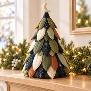 May include: A decorative cone-shaped tree ornament crafted from layered fabric leaves in shades of navy blue, olive green, cream, and rust orange. The leaves feature gold embroidered star and branch patterns, topped with a small cream crescent moon.