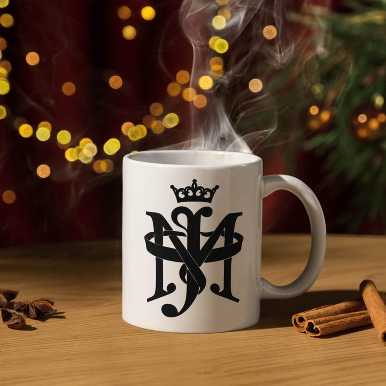 May include: A white ceramic mug with a black monogram design featuring the letters "M", "S", and "J" intertwined, topped with a crown. Steam rises from the mug, suggesting a hot beverage. The mug sits on a wooden surface.