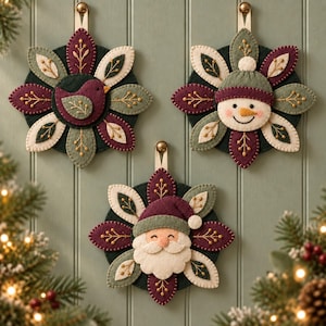 May include: Three circular felt ornaments featuring a bird, a snowman, and Santa Claus. Each ornament has layered petal-shaped felt pieces in dark green, cream, and deep plum with gold embroidered branch details and a hanging loop at the top.