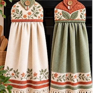 May include: Two hanging kitchen towels with waffle-weave fabric, one cream and one sage green. Both feature a floral print border with orange flowers and green leaves, a quilted top tab with a wooden button, and a cream-colored fringe trim at the bottom.