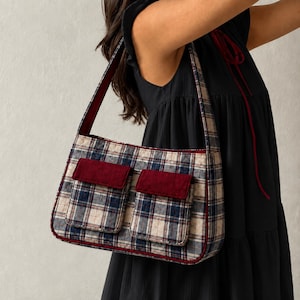 May include: A rectangular shoulder bag featuring a blue, white, and black plaid pattern with two front pockets. The pockets have solid dark red flaps, and the bag is finished with dark red piping along the edges and a matching shoulder strap.