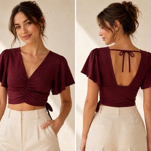 Op de afbeelding: Een diepbordeauxrode crop top met korte vlindermouwen, een V-hals en een geplooid detail aan de voorzijde. Het rugontwerp heeft een diepe open uitsnijding met een dunne striksluiting aan de bovenkant, gecombineerd met een crèmekleurige broek.
