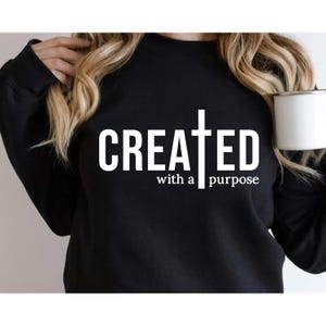 May include: A black long-sleeve sweatshirt featuring white text that reads CREATED with a purpose, where the letter T is replaced by a vertical cross symbol. The garment has a crew neckline and long sleeves, providing a simple and clean design.