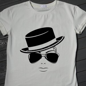 May include: A white t-shirt featuring a black and white graphic of a face wearing a fedora hat and sunglasses. The design is a minimalist illustration, with clean lines and a modern aesthetic. The shirt is made of a soft, comfortable material.
